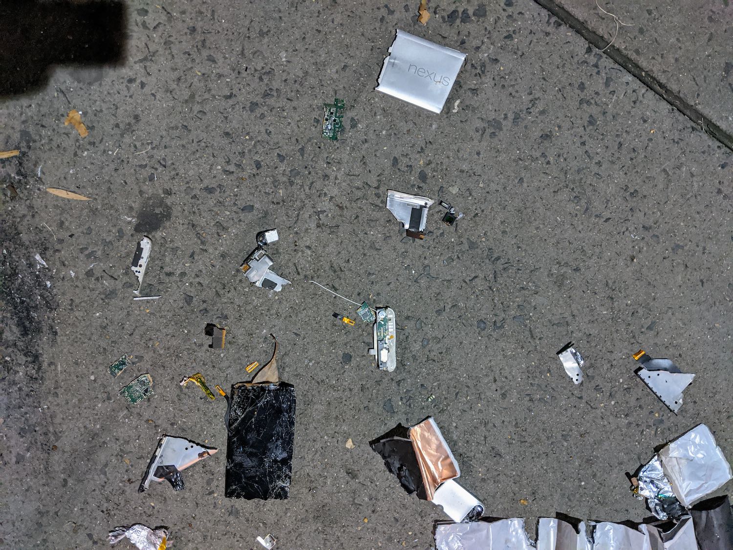 A Nexus 6P phone lays smashed to smithereens on a concrete pavement. Green pieces of electronic component board are smashed up and scattered around. The phone screen appears more like the surface of a cheese grater.