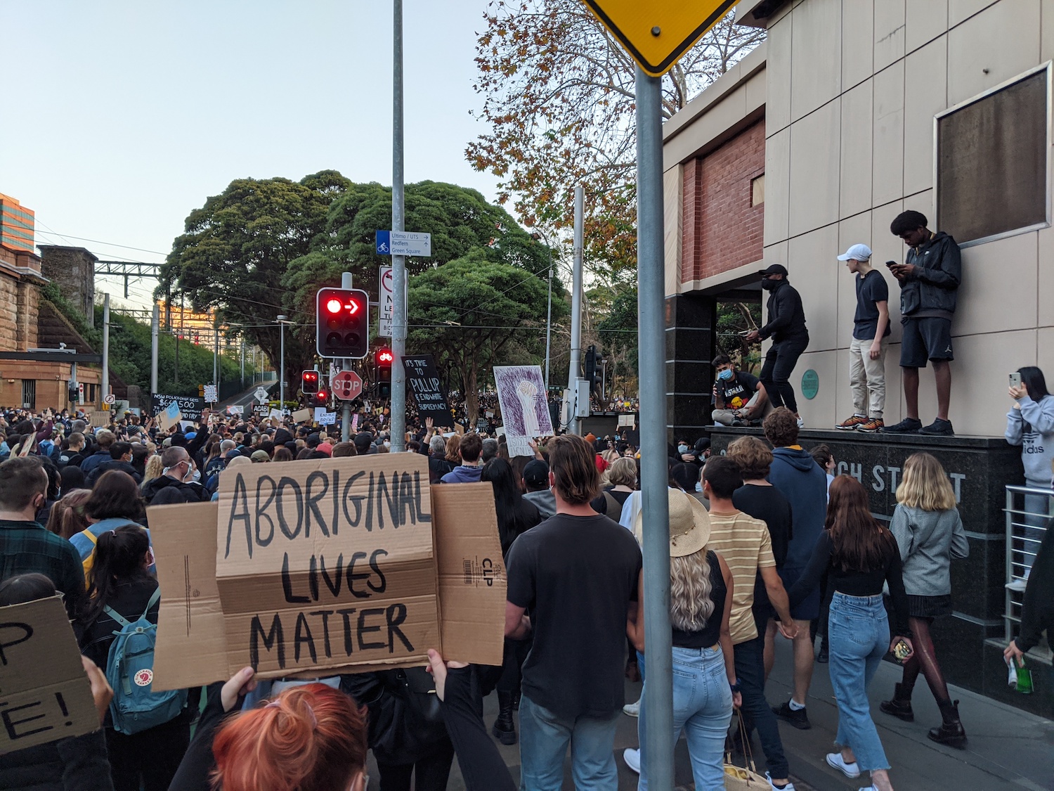 Protesters marching around a corner in to Belmore Park. A protestor is holding a cardboard sign saying "Aboriginal Lives Matter"